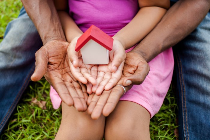 Child sitting on adults lap, both holding out hands carrying a model house